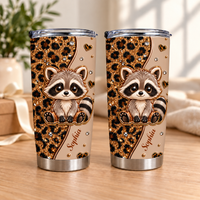 Two stainless steel tumblers with a cute raccoon and leopard print design stand upright on a wooden table near a window.