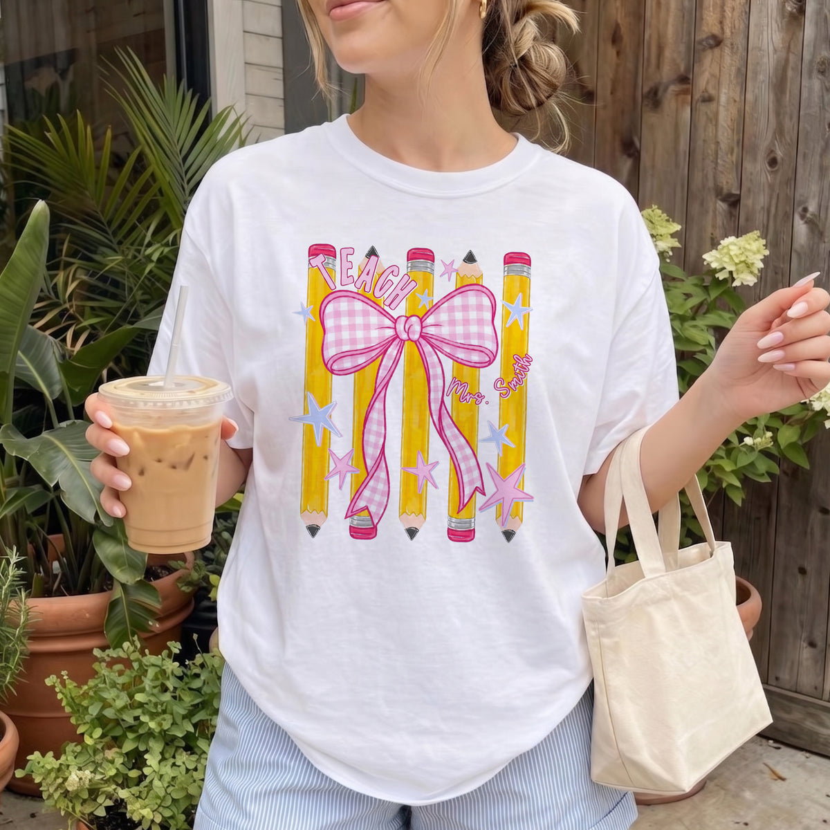 A white t-shirt featuring a pink bow and pencil graphic, worn by a person holding an iced coffee next to potted plants.