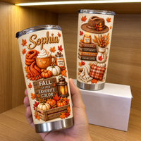 A hand holds a custom name stainless steel tumbler with autumn designs in front of a shelf with another cup.