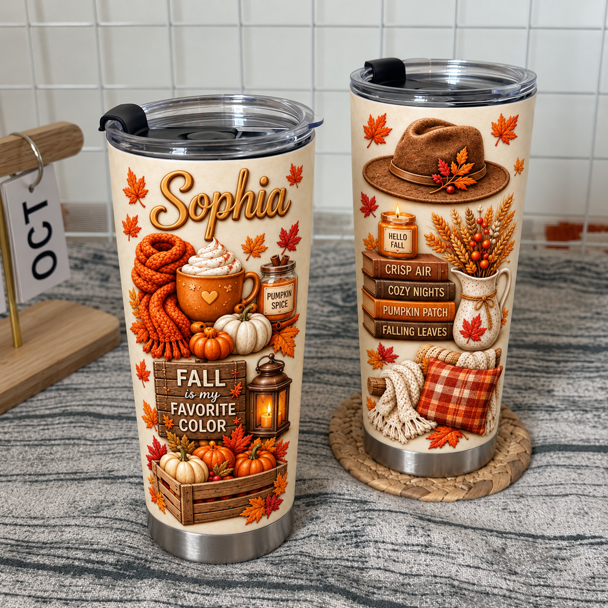 Two custom name hello fall tumblers with autumn graphics stand on a textured surface next to a calendar.