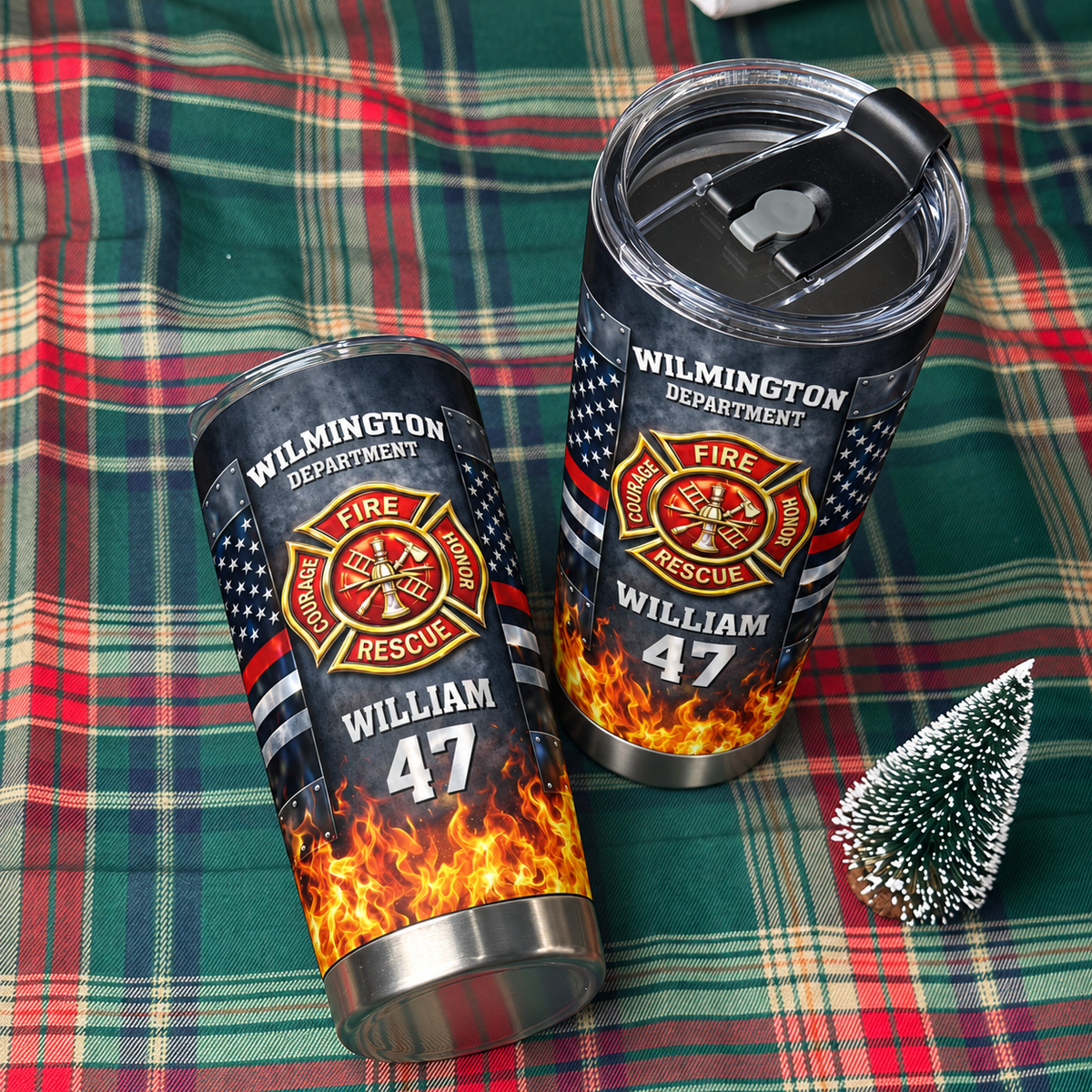 Two personalized firefighter tumblers displayed on a green and red plaid fabric next to a miniature Christmas tree.