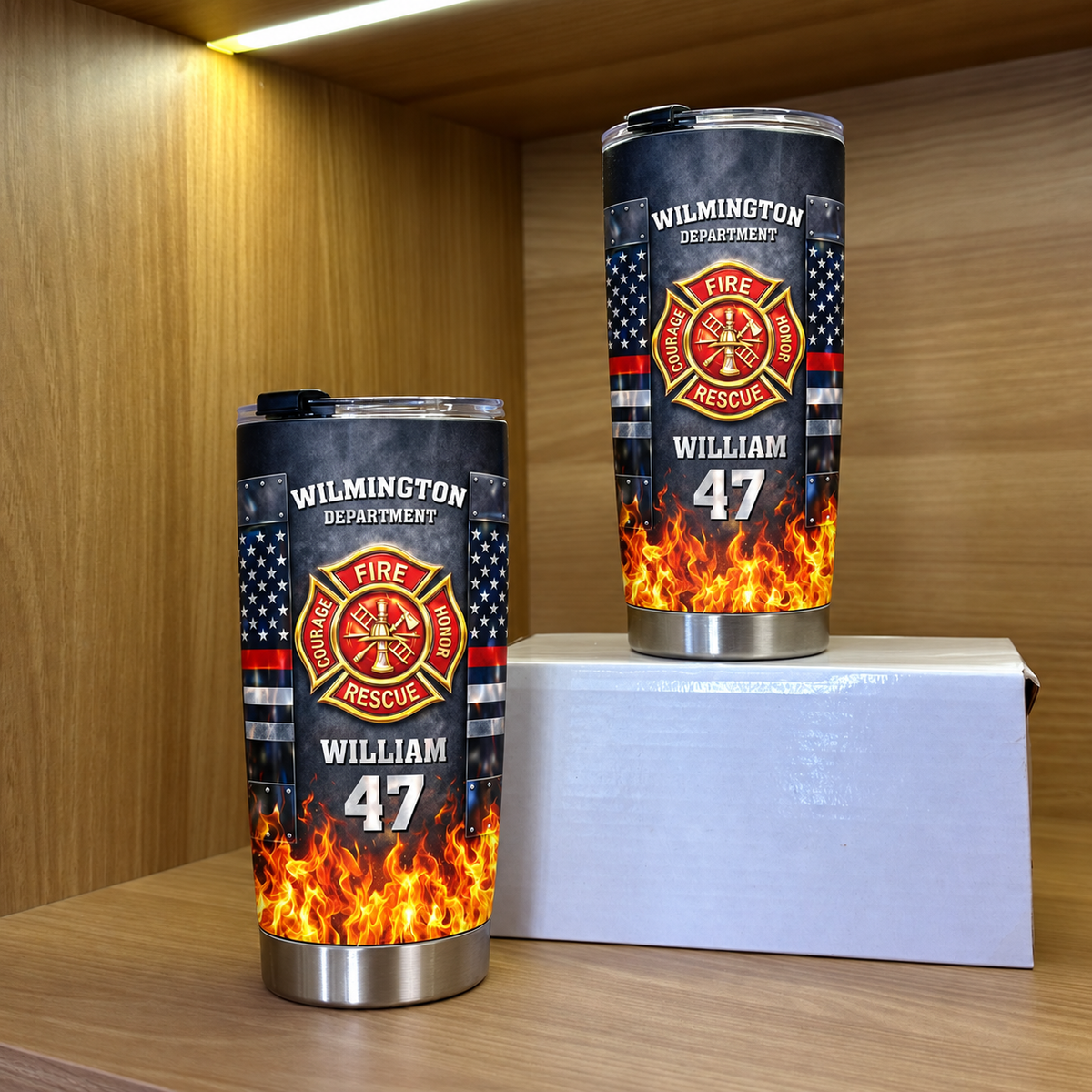 Two personalized stainless steel travel mugs with firefighter badge design displayed on a white box inside a wooden cabinet.