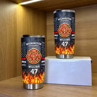 Two personalized stainless steel travel mugs with firefighter badge design displayed on a white box inside a wooden cabinet.