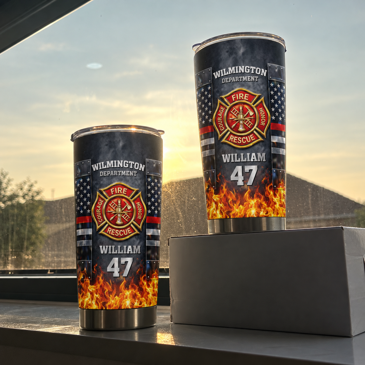 Two personalized stainless steel firefighter tumblers standing on a box near a window at sunset with soft outdoor light.