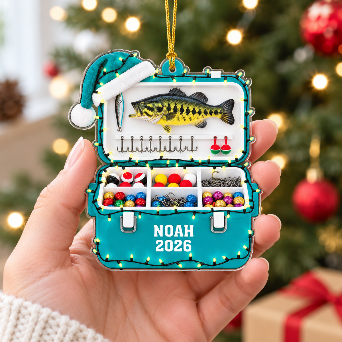 Hand holding a teal personalized fishing gift acrylic ornament featuring a tackle box design, bass fish, and NOAH 2026 text.
