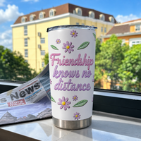 A floral stainless steel cup with the friendship quote standing on a windowsill with a blurry building background and