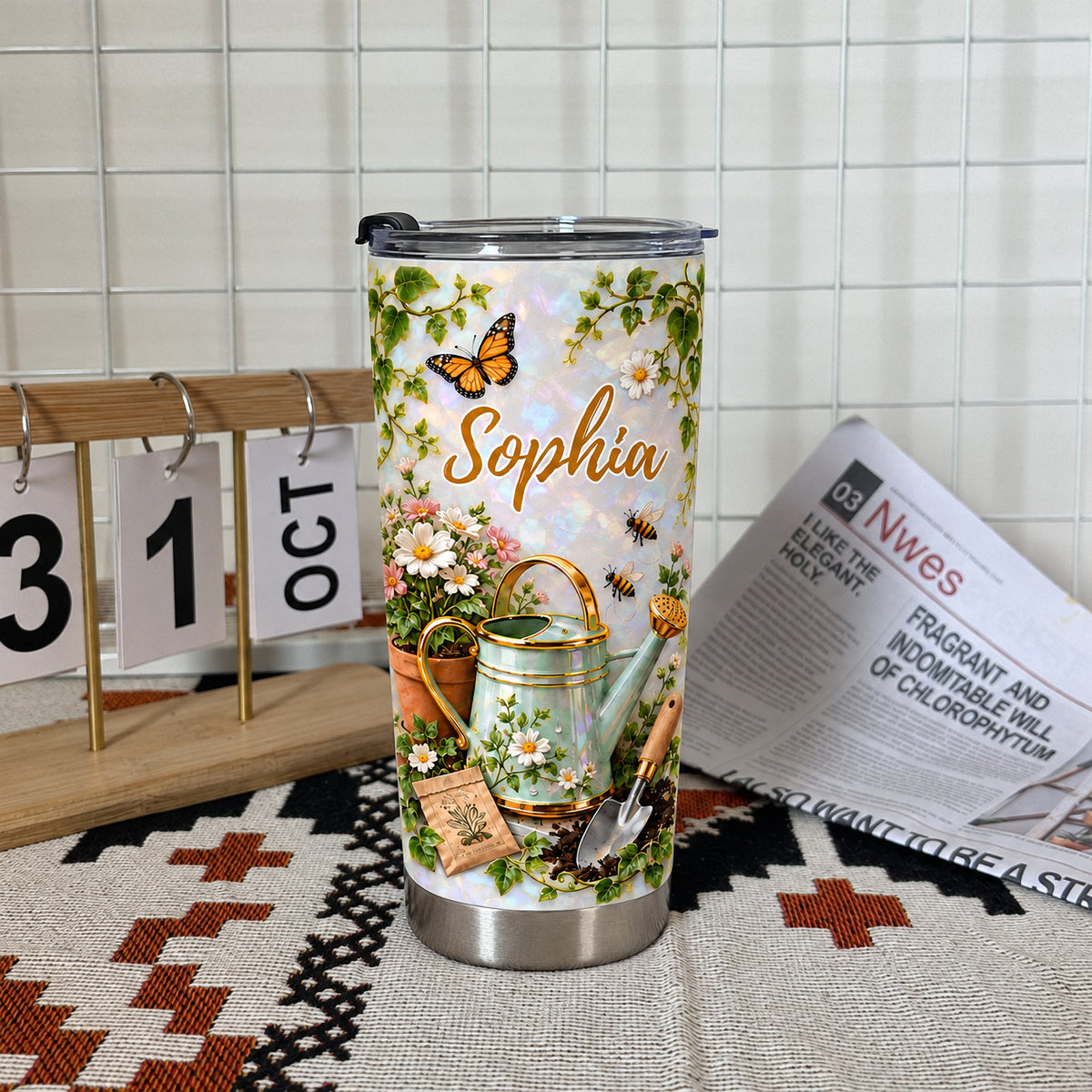 Gardening theme personalized tumbler with butterfly and watering can illustration standing upright on a patterned cloth