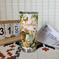 Gardening theme personalized tumbler with butterfly and watering can illustration standing upright on a patterned cloth