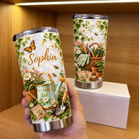 Hand holding a personalized gardening tumbler next to a second tumbler showcasing garden basket artwork on a wooden shelf.