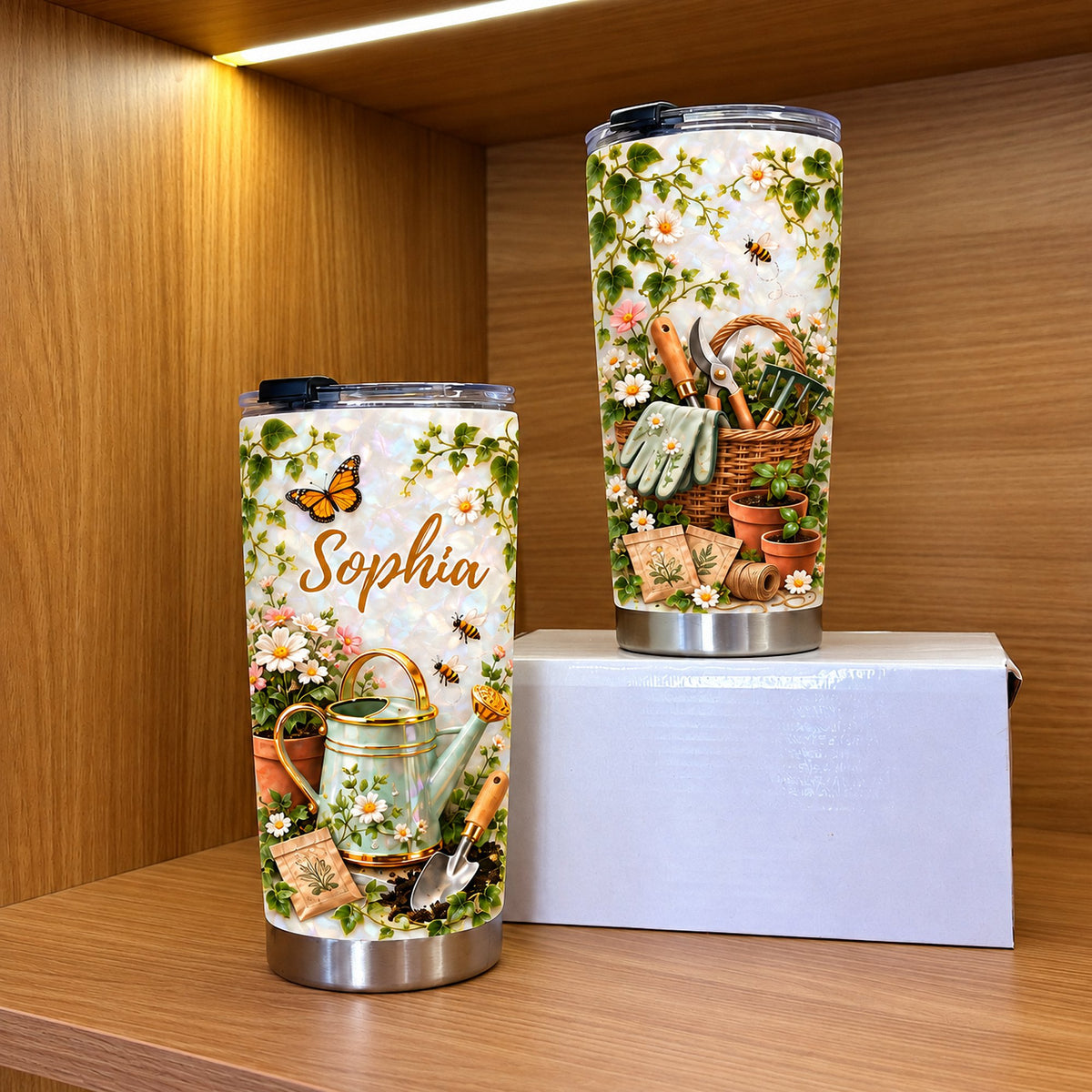 Two personalized botanical tumblers with floral and gardening designs standing on a white box inside a wooden display shelf.