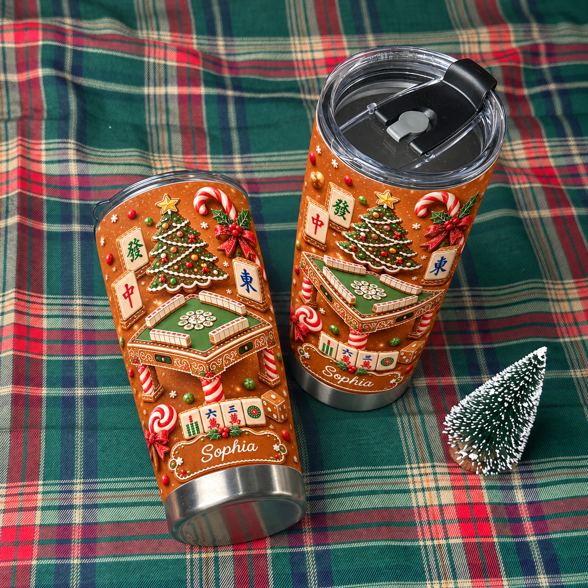 Two stainless steel tumblers with gingerbread mahjong design resting on a red and green plaid cloth with mini tree.