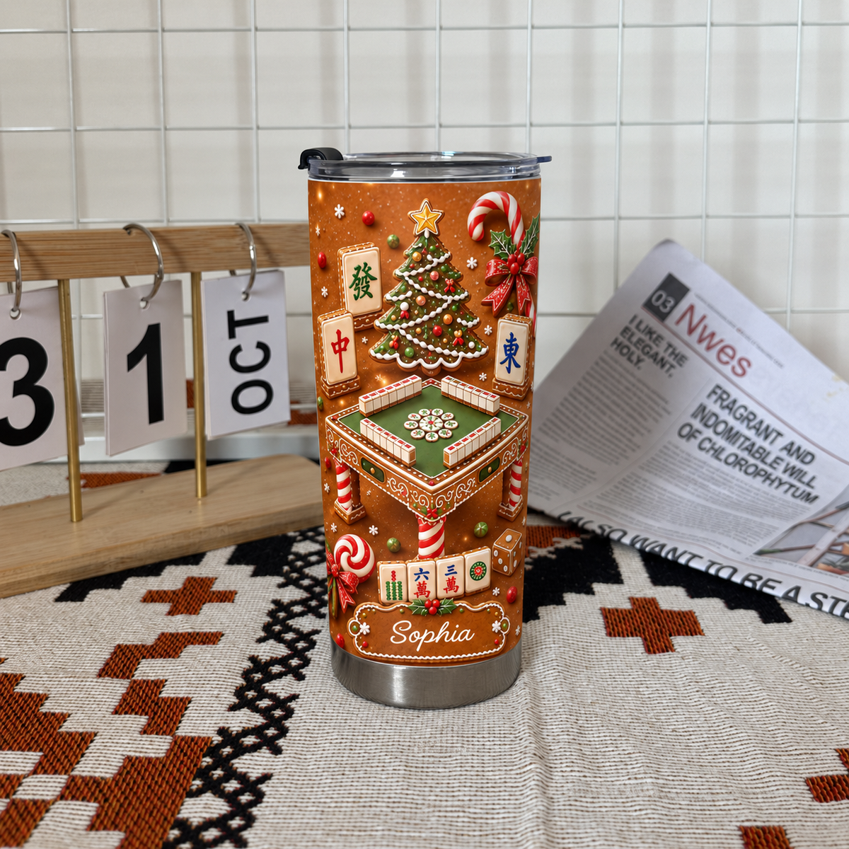 A custom name tumbler featuring gingerbread mahjong design next to a wooden perpetual calendar and newspaper.