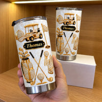 A hand holds a gold diamond golf theme tumbler next to a white box on a wooden shelf.