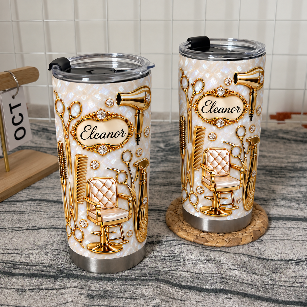 Two stainless steel travel mugs with gold hairdresser tools designs displayed on textured surfaces with a calendar.