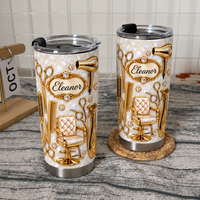 Two stainless steel travel mugs with gold hairdresser tools designs displayed on textured surfaces with a calendar.