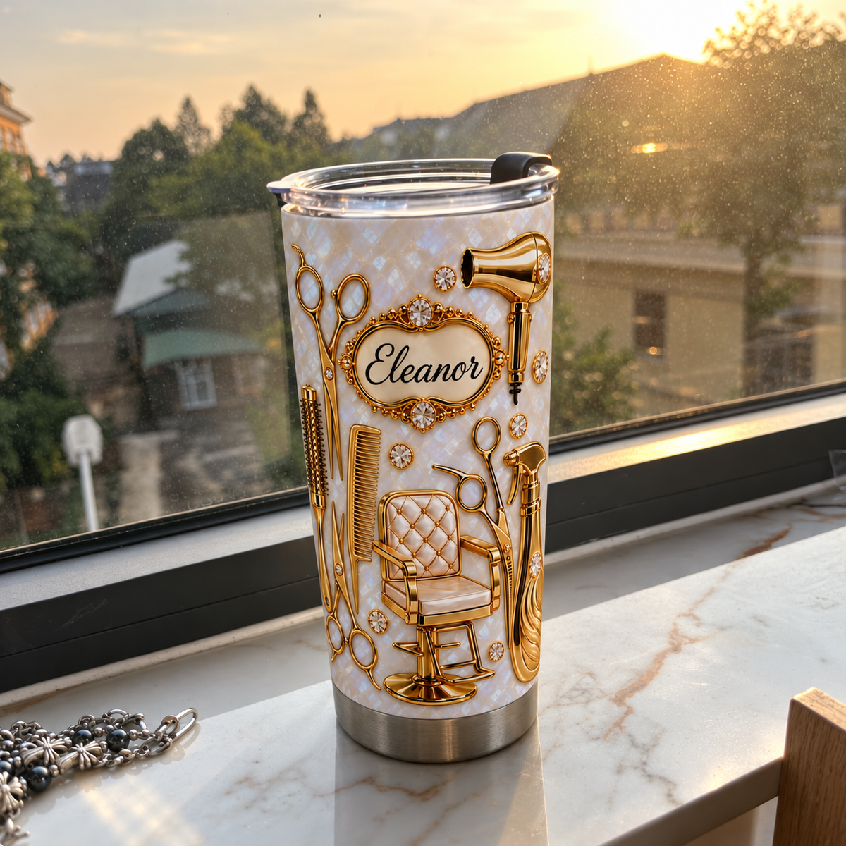 Stainless steel travel mug with gold salon tools pattern resting on a white marble windowsill at sunset.