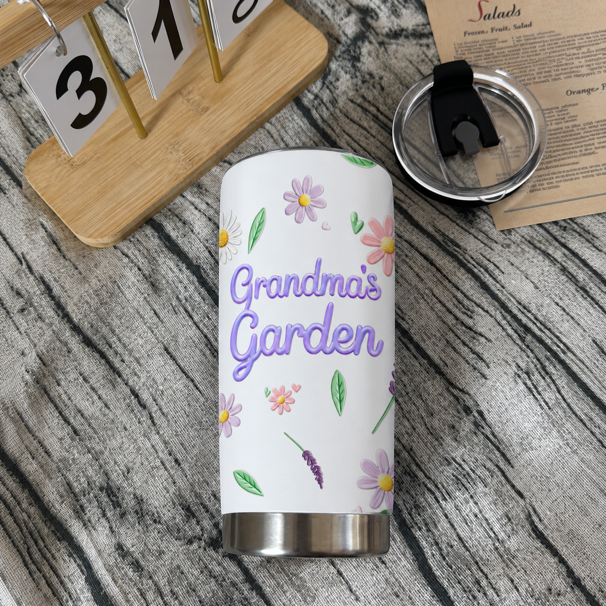 A custom birth flower tumbler lying horizontally on a wooden surface beside a desk calendar.