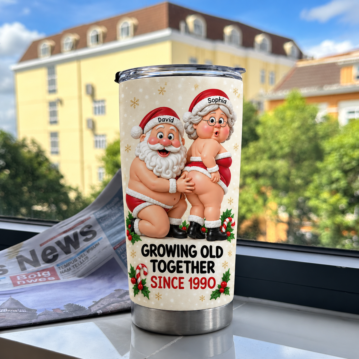 Personalized anniversary gift tumbler displaying naughty Santa graphic beside a newspaper and buildings.