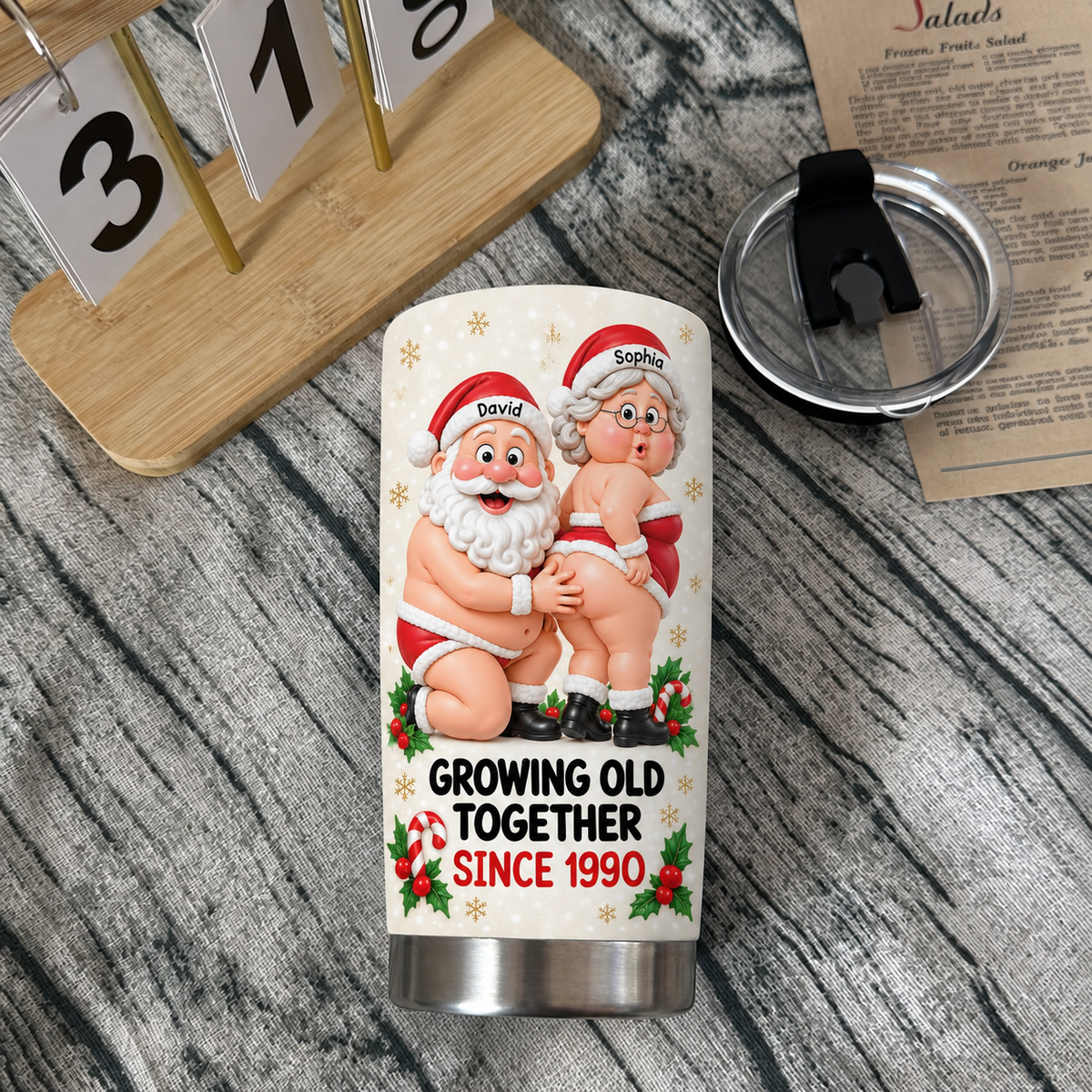 Stainless steel tumbler with festive Santa graphic resting on textured wood table next to open lid and calendar.