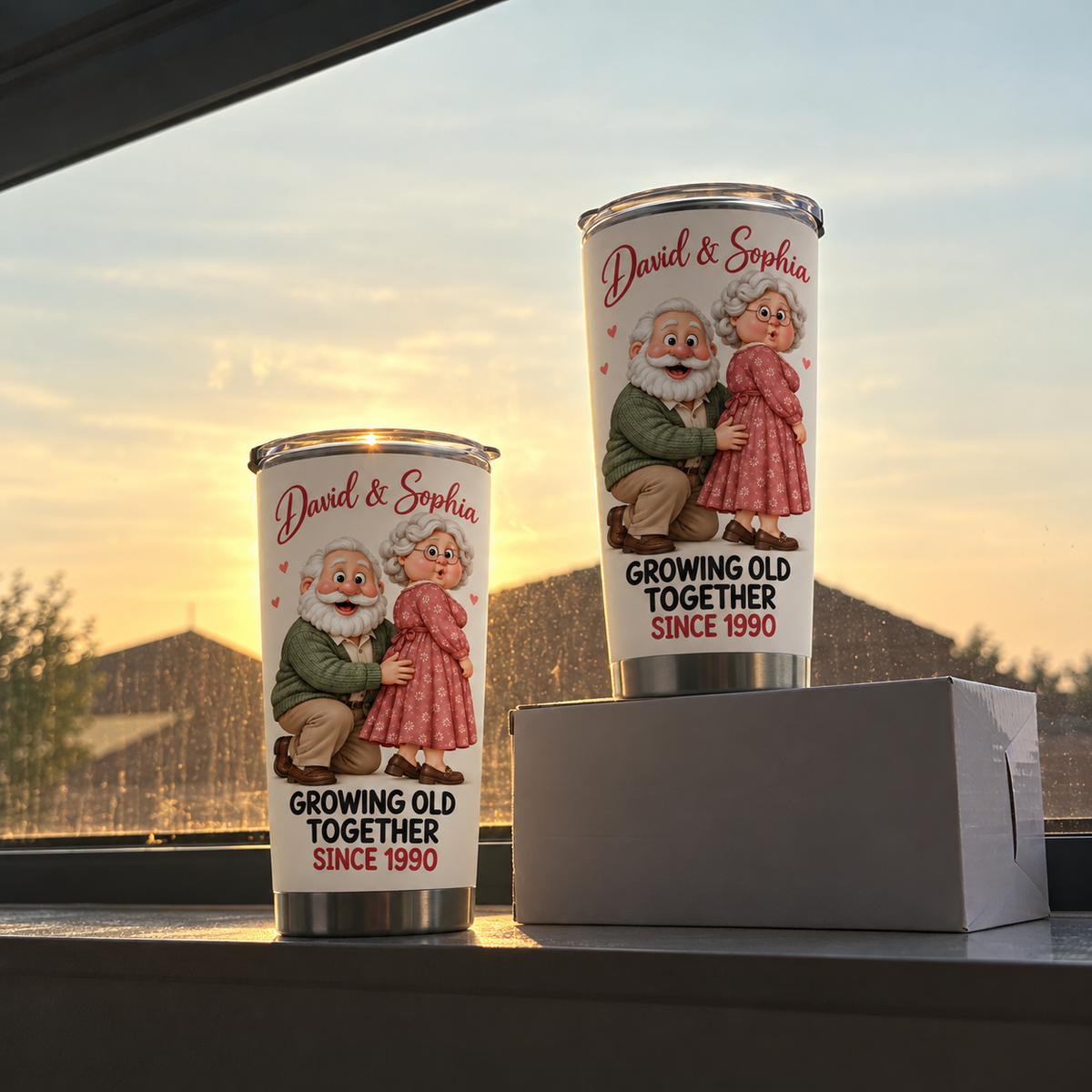 Two white stainless steel travel tumblers with cartoon elderly couple design standing on a white box against a window view at