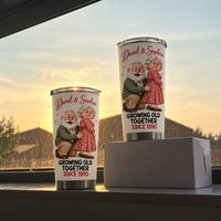 Two white stainless steel travel tumblers with cartoon elderly couple design standing on a white box against a window view at