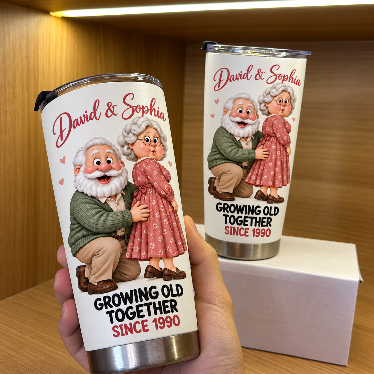 Hand holding a white stainless steel travel tumbler with an illustrated elderly couple and David & Sophia text, next to a