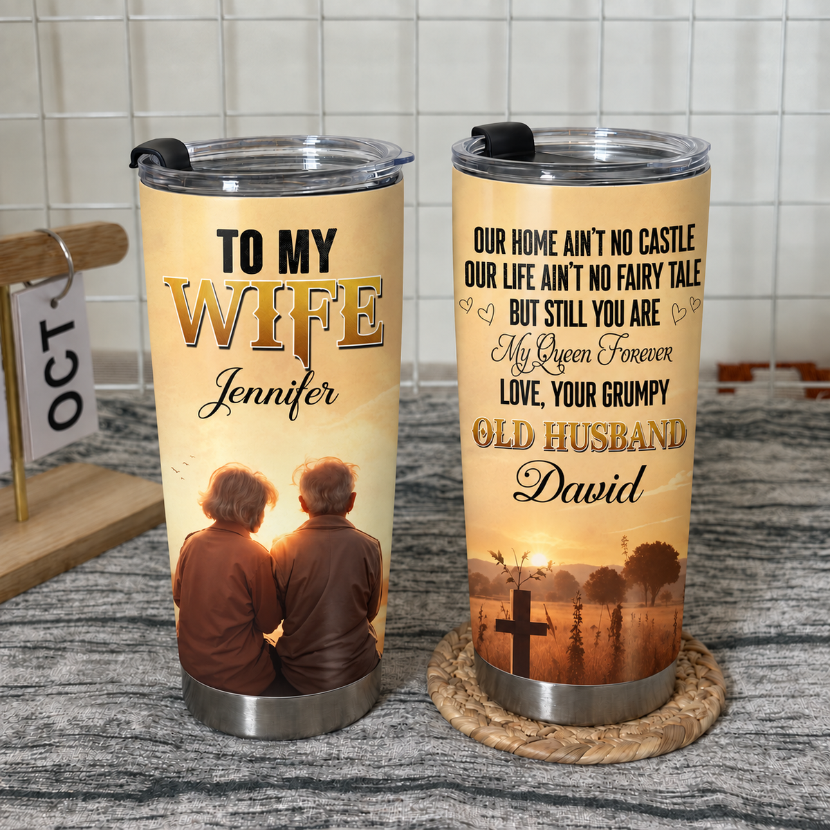 Two stainless steel travel mugs showing front and back designs resting on a woven coaster on a textured surface.