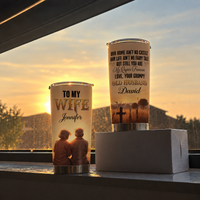Two 20oz stainless steel travel mugs with sunset couple graphics displayed on a windowsill against a sunset sky.