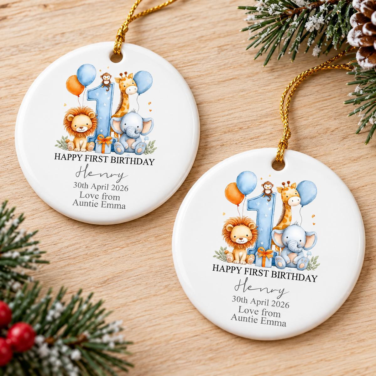 Two personalized baby gift ceramic ornaments resting side by side on a wooden table with pine greenery.