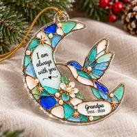 Blue hummingbird memorial ornament with custom text resting on a light neutral cloth surrounded by pine needles and berries.