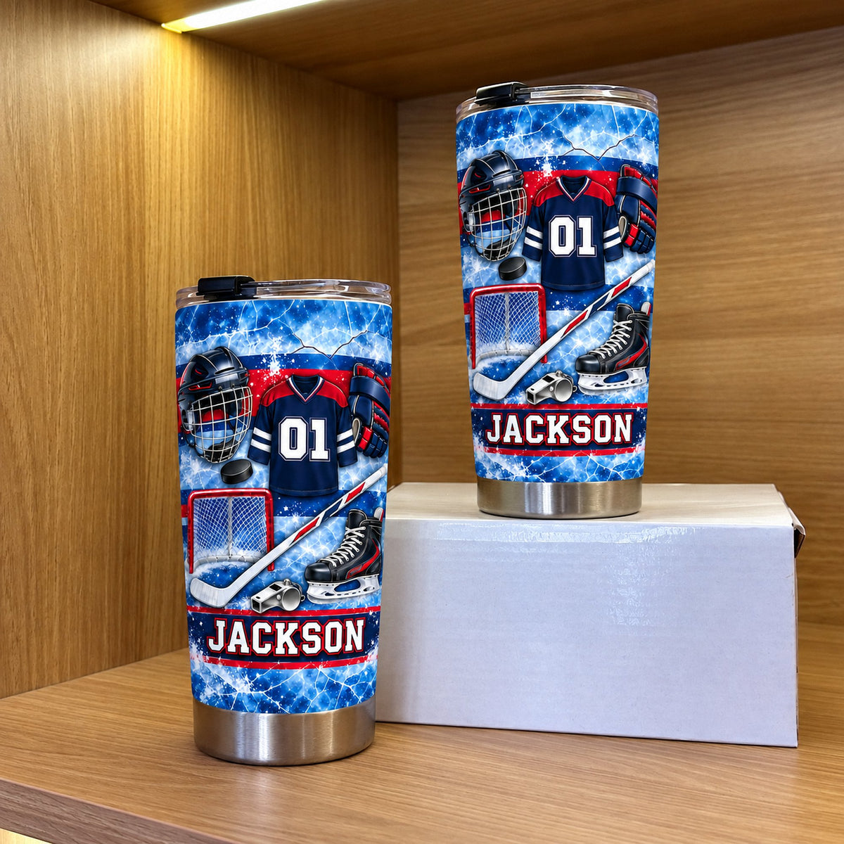 Two personalized hockey gear tumblers of different sizes displayed on a white box inside a wooden cabinet shelf.