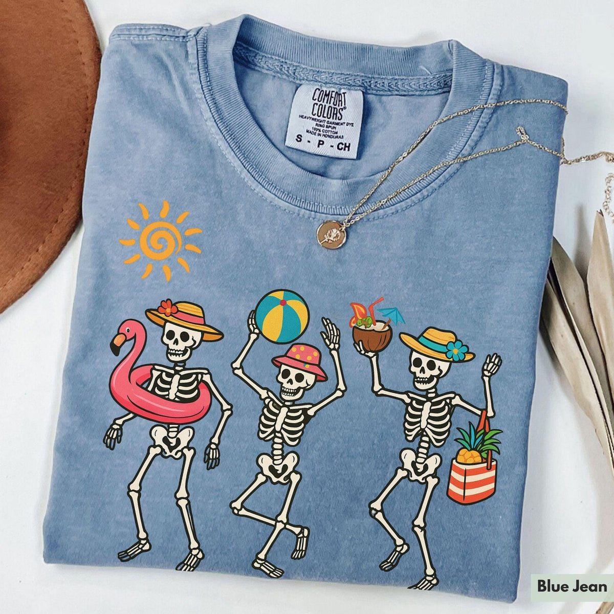 Blue-grey graphic t-shirt featuring three dancing skeletons with beach accessories like a flamingo float, beach ball, and