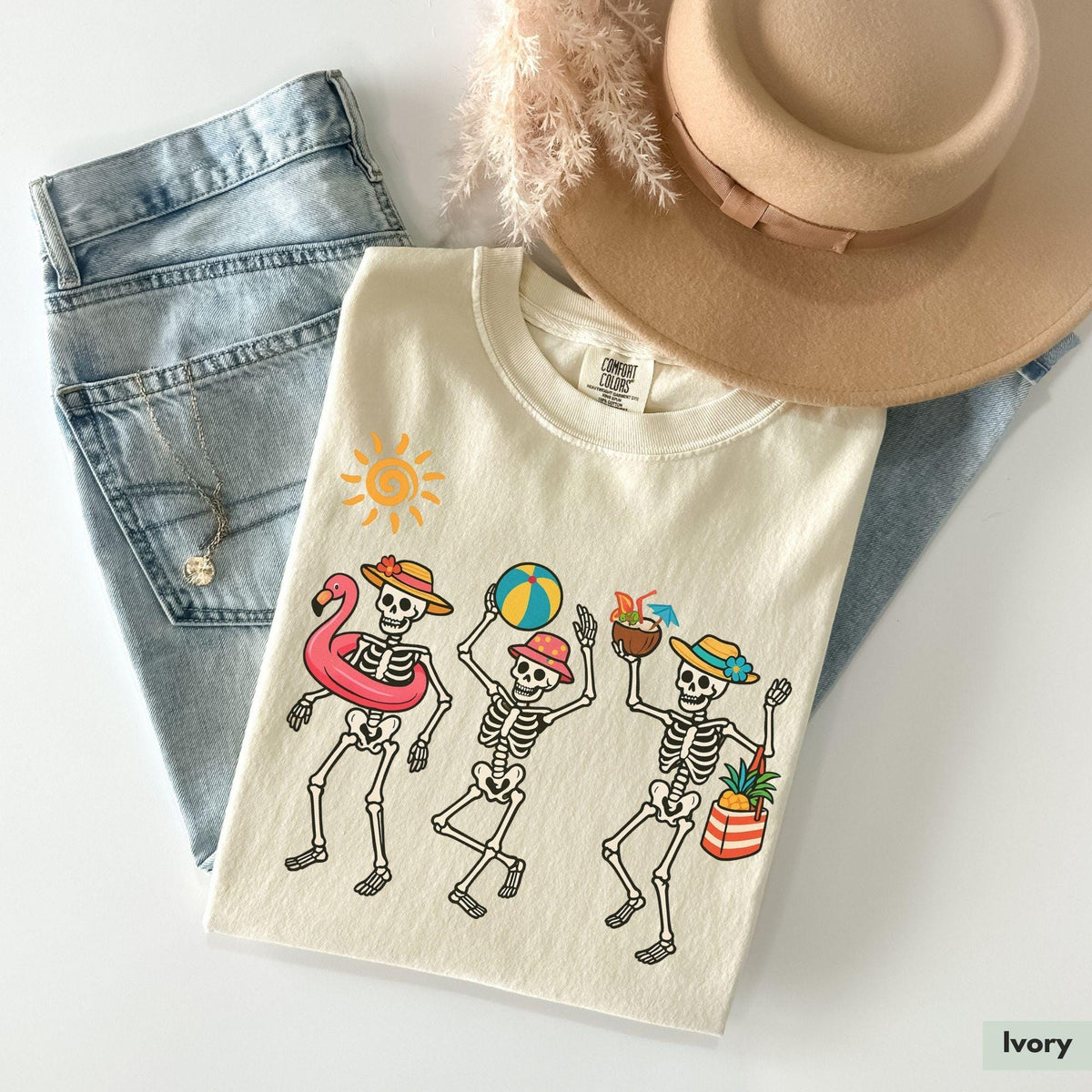 Ivory t-shirt featuring a beach skeleton graphic, laid flat next to jeans and a hat.