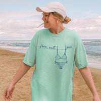 A woman in a mint green t-shirt with a blue bikini and clothesline graphic, standing on a sandy beach.