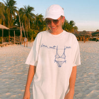 A woman wearing an off-white t-shirt with a blue bikini graphic, standing on a sunny beach with palms.