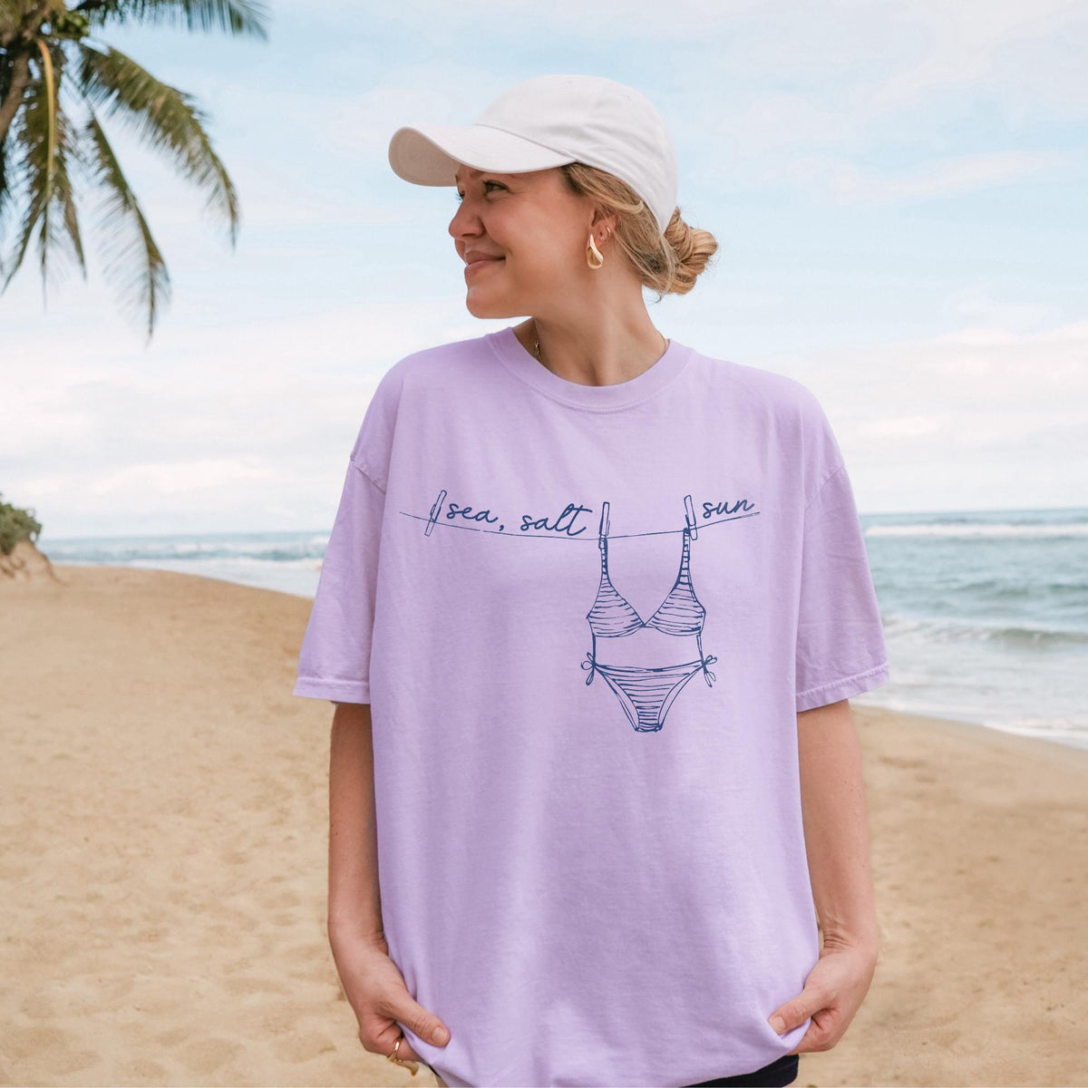 A woman wearing a light purple t-shirt with a blue bikini and clothesline graphic, posing on a beach.