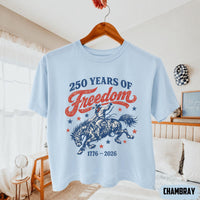 A light blue t-shirt hanging on a wooden hanger showing a vintage Western 250 Years of Freedom graphic.
