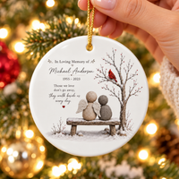 Hand holding the round white ceramic memorial ornament featuring a pebble angel design and gold string.