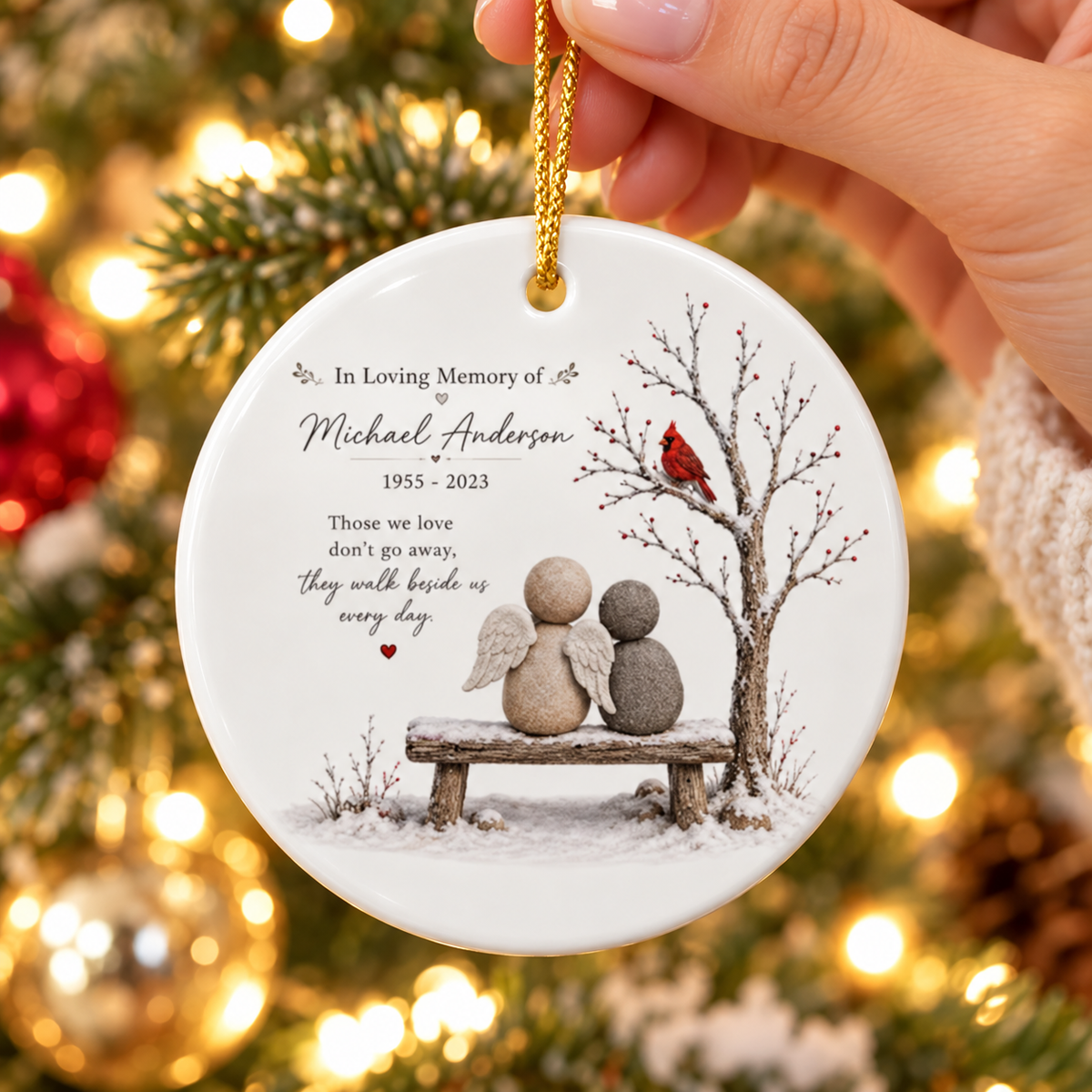 Hand holding the round white ceramic memorial ornament featuring a pebble angel design and gold string.