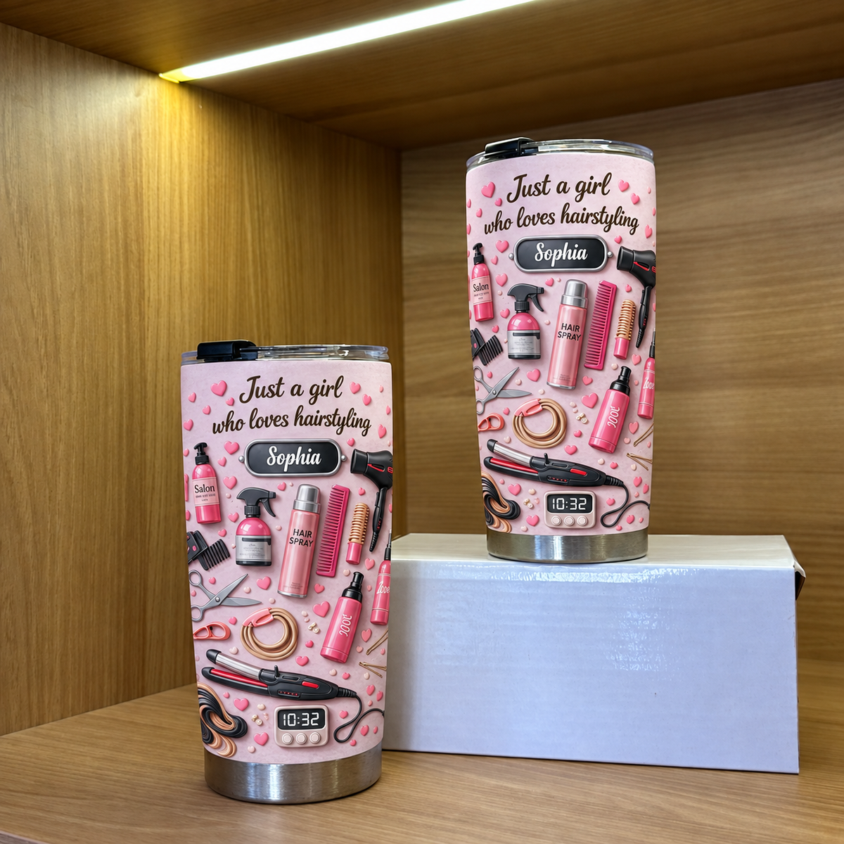 Two personalized hair stylist tumblers in pink on a wooden shelf with warm overhead lighting.