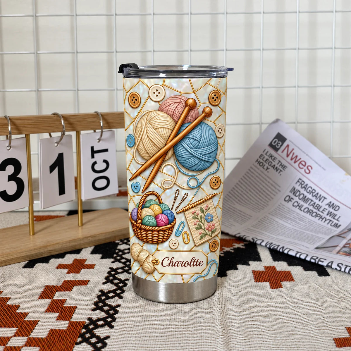 Insulated stainless steel tumbler with knitting yarn design standing upright on a textured mat next to a desk calendar and