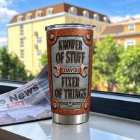 A custom handyman tumbler sits on a windowsill next to a newspaper with blurred buildings and trees outside.