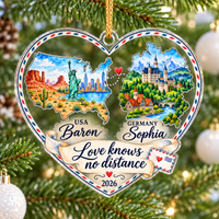 Personalized long distance relationship ornament hanging among pine needles with soft holiday lights bokeh in the background.