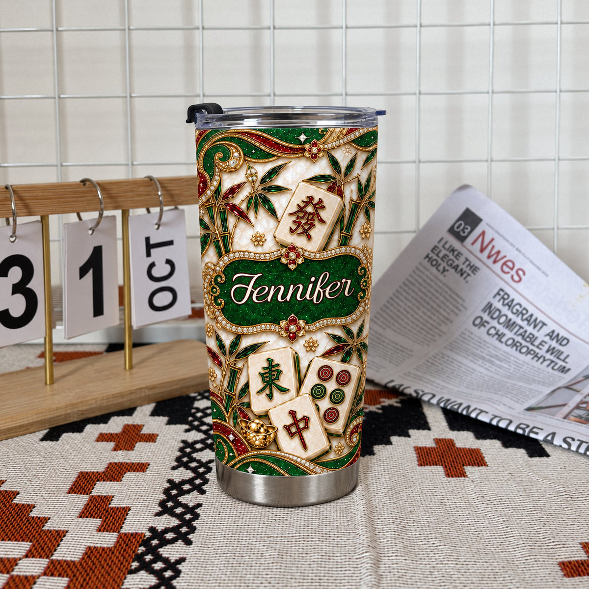 Personalized mahjong tumbler on patterned mat next to small calendar block and newspaper in bright room.