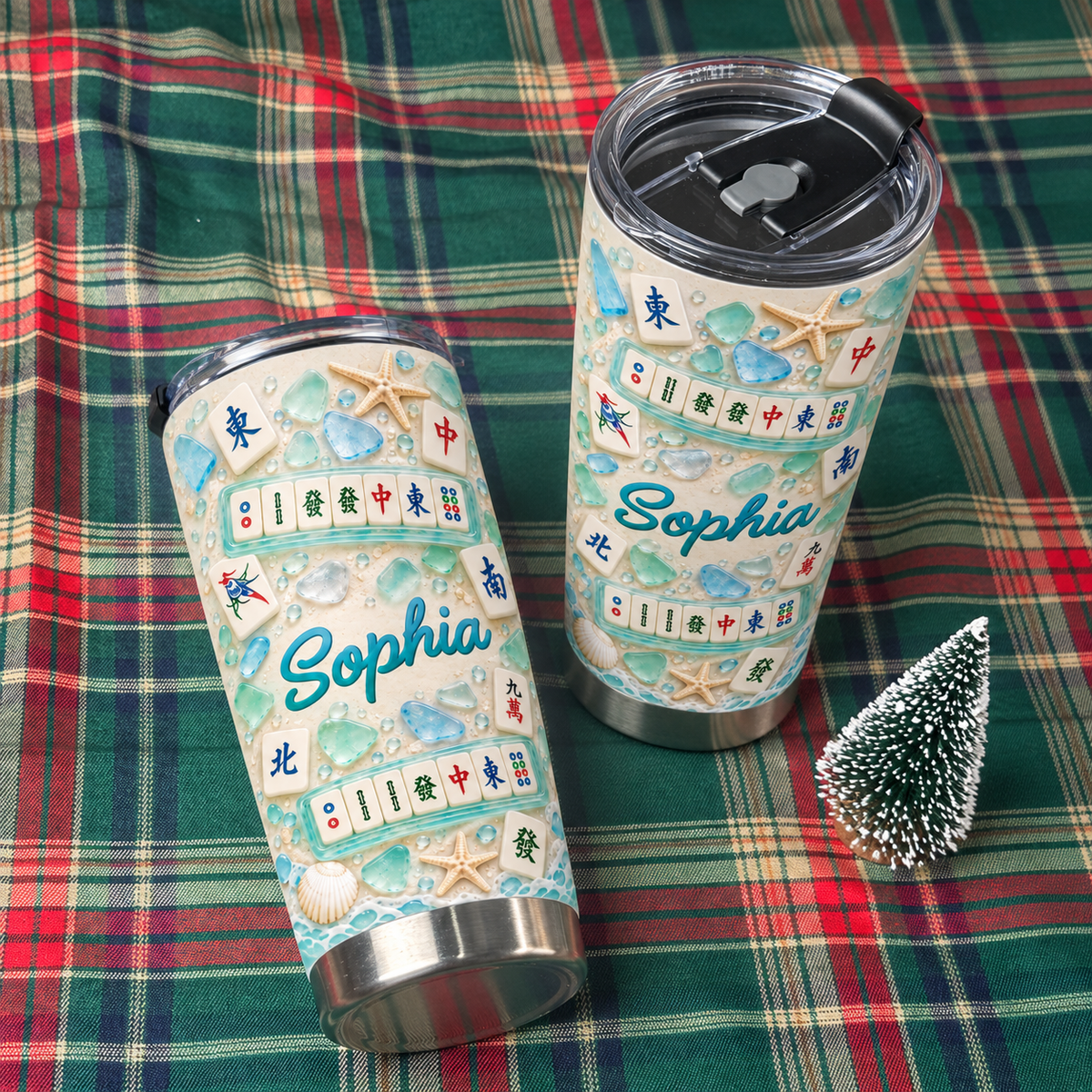 A custom name tumbler lies on a red and green plaid blanket beside a tiny pine tree.