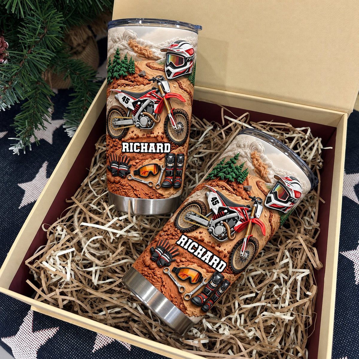 Two personalized dirt bike rider cups in open gift box lined with filler and pine branch in background.