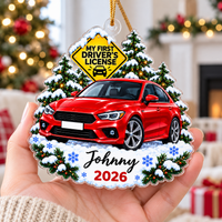 Hand holding a personalized new driver acrylic ornament with a red car and snowy pine trees in front of a blurred holiday
