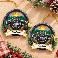 Two personalized new driver acrylic ornaments laid flat on a wooden surface next to holiday pine branches.