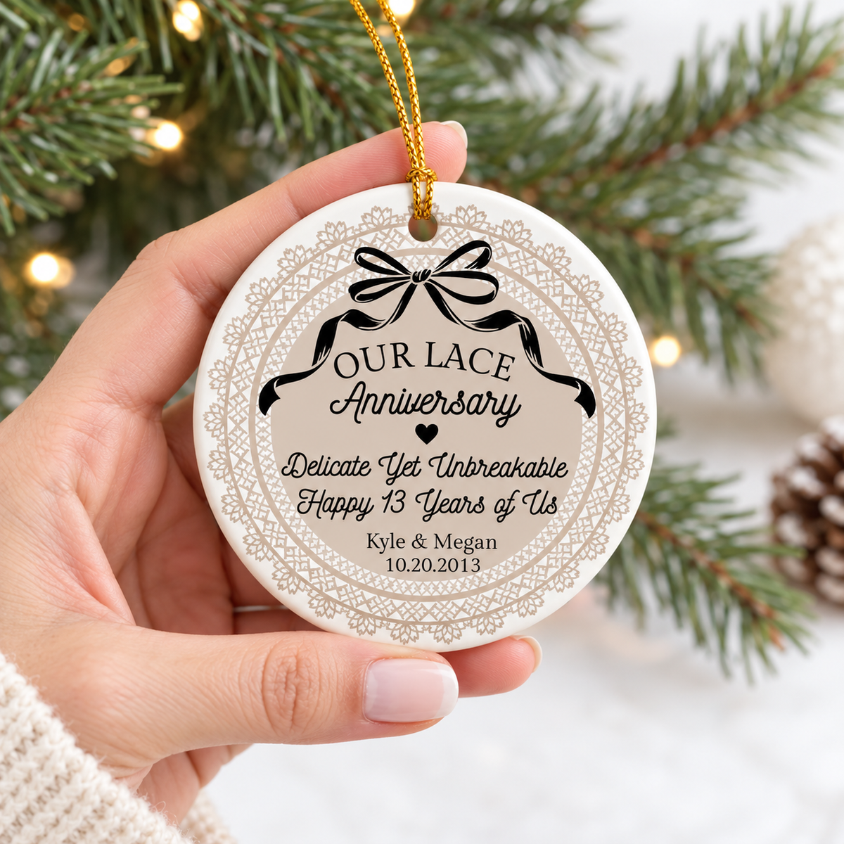 Hand holding a personalized round ceramic lace anniversary ornament with a beige lace pattern and gold string against holiday