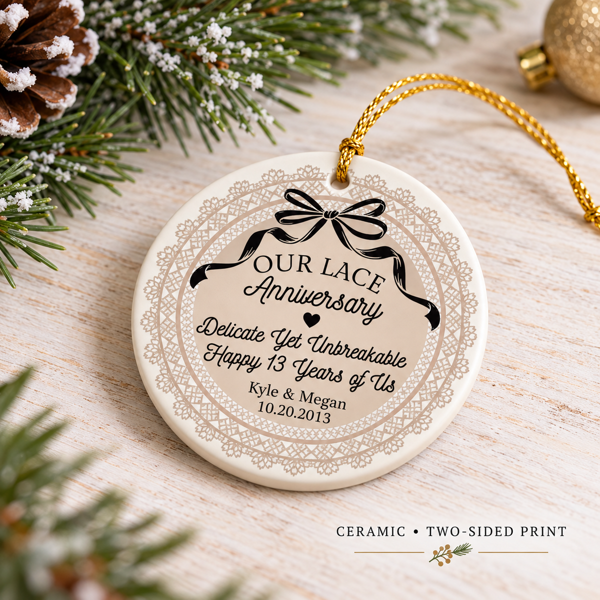 Round ceramic lace anniversary ornament lying on a wooden table next to pine cones and a gold ornament ball with text